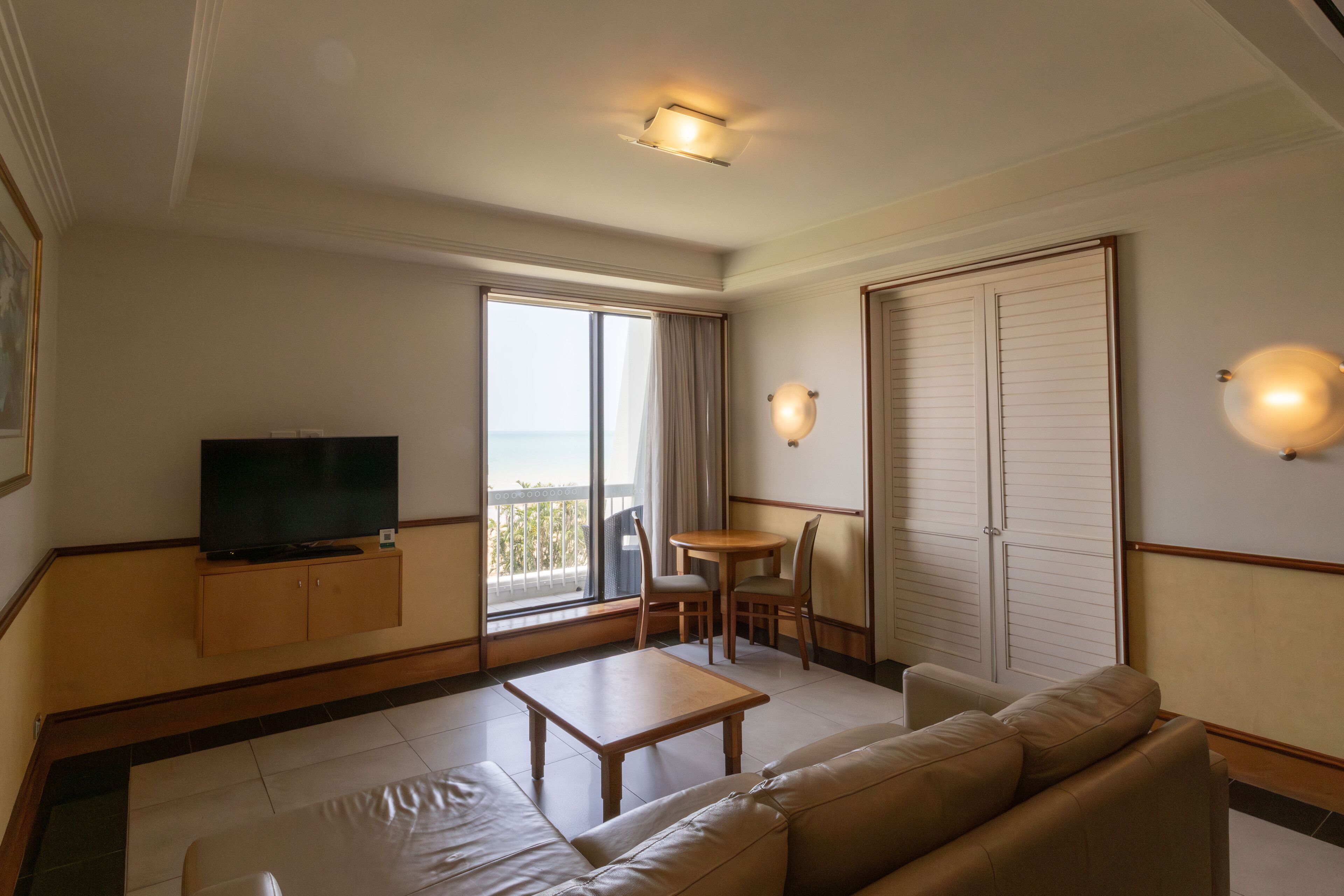 Quarto de hotel com sofá, uma pequena mesa, televisão, varanda com vista e guarda-roupa.