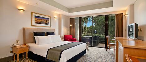 A hotel room with a large bed, a desk, a chair, a TV, and a balcony with a view of trees.