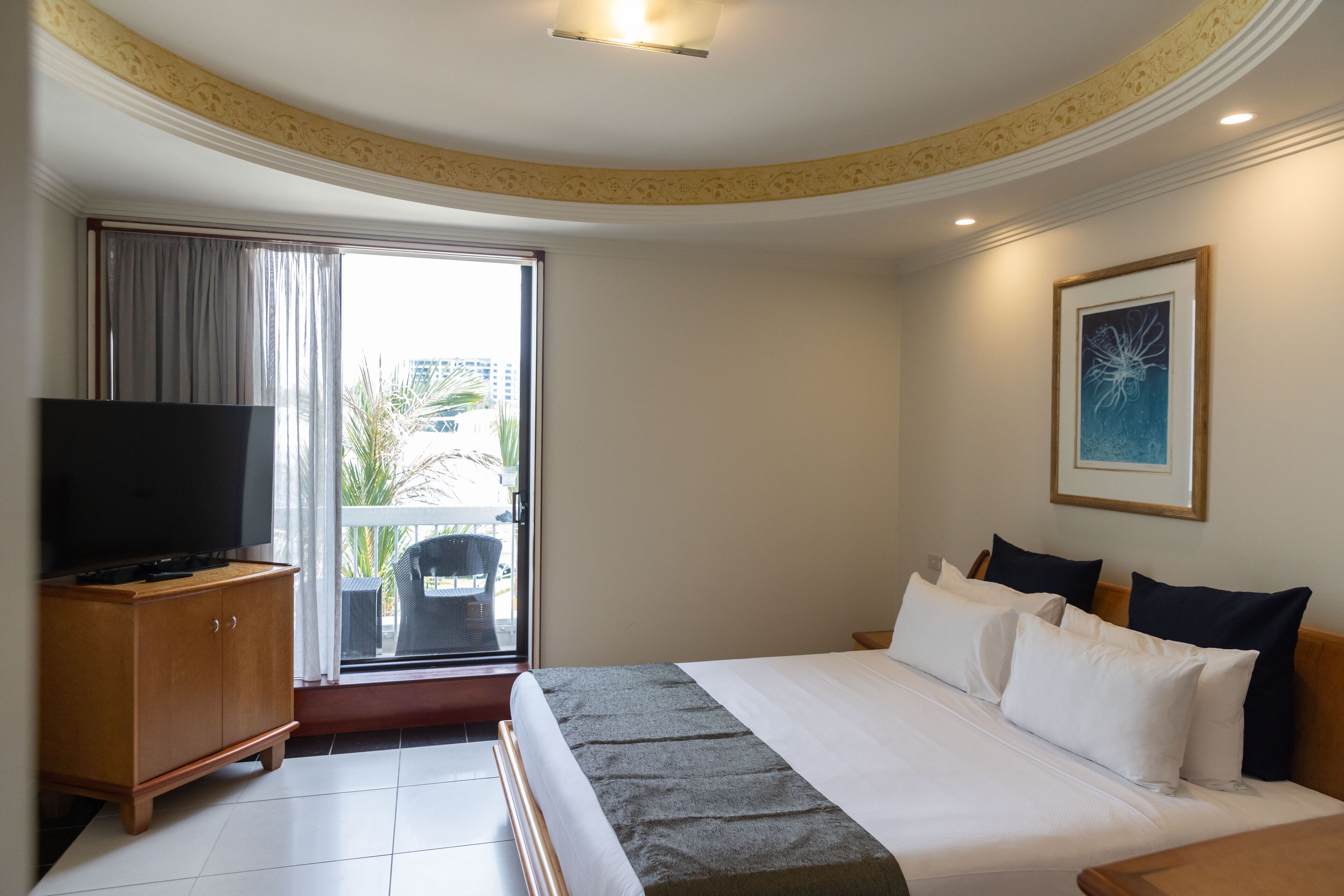A hotel room with a bed, a TV on a wooden cabinet, a balcony with a view of palm trees, and a framed artwork on the wall.