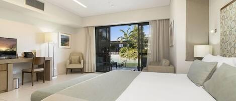 A modern hotel room with a large bed, a desk, a chair, and a balcony with a view of palm trees.
