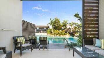 Un espacio moderno al aire libre con mobiliario de mimbre y una mesa de cristal, con vistas a una piscina y palmeras.