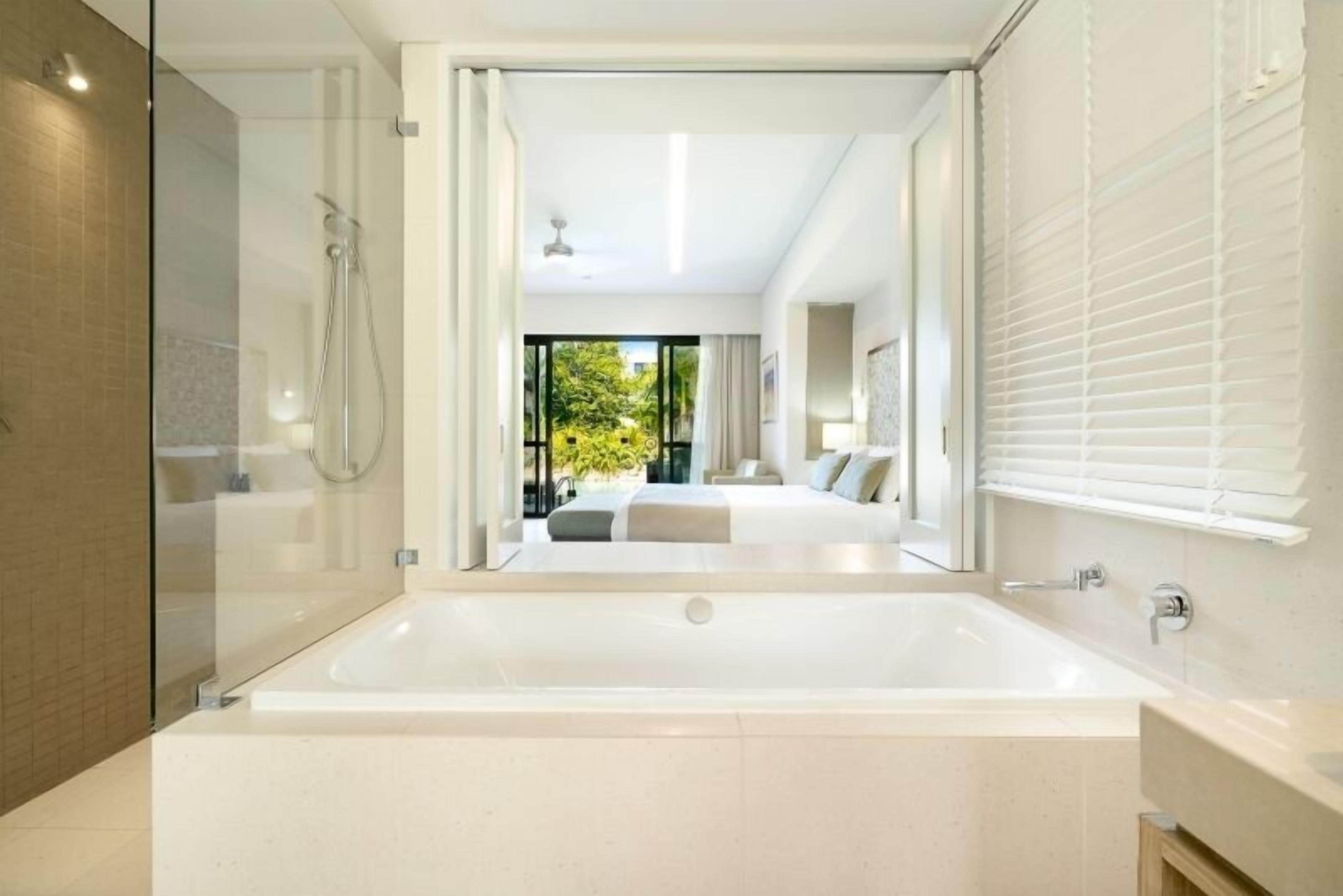 Een moderne badkamer met een ruime badkuip, een douchecabine van glas en uitzicht op een slaapkamer met een bed en balkon.