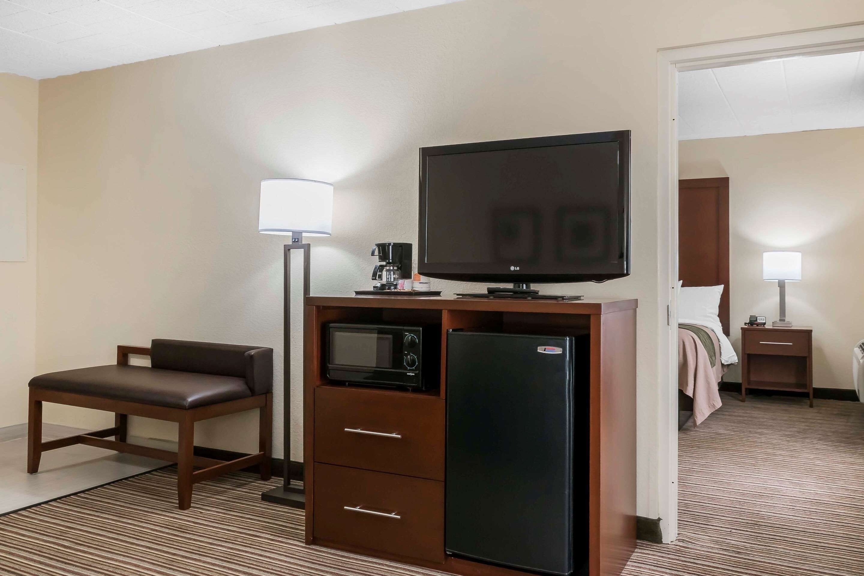 A compact hotel room with a kitchenette, a flat-screen TV, a microwave, a refrigerator, a sofa, and a bedside table with a lamp.
