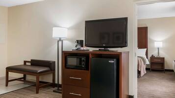 A compact hotel room with a kitchenette, a flat-screen TV, a microwave, a refrigerator, a sofa, and a bedside table with a lamp.