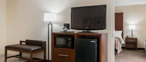 A compact hotel room with a kitchenette, a flat-screen TV, a microwave, a refrigerator, a sofa, and a bedside table with a lamp.