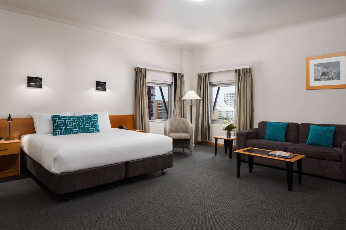 Habitación de hotel con cama, sofá, sillón, una mesita de centro y dos ventanas con cortinas.