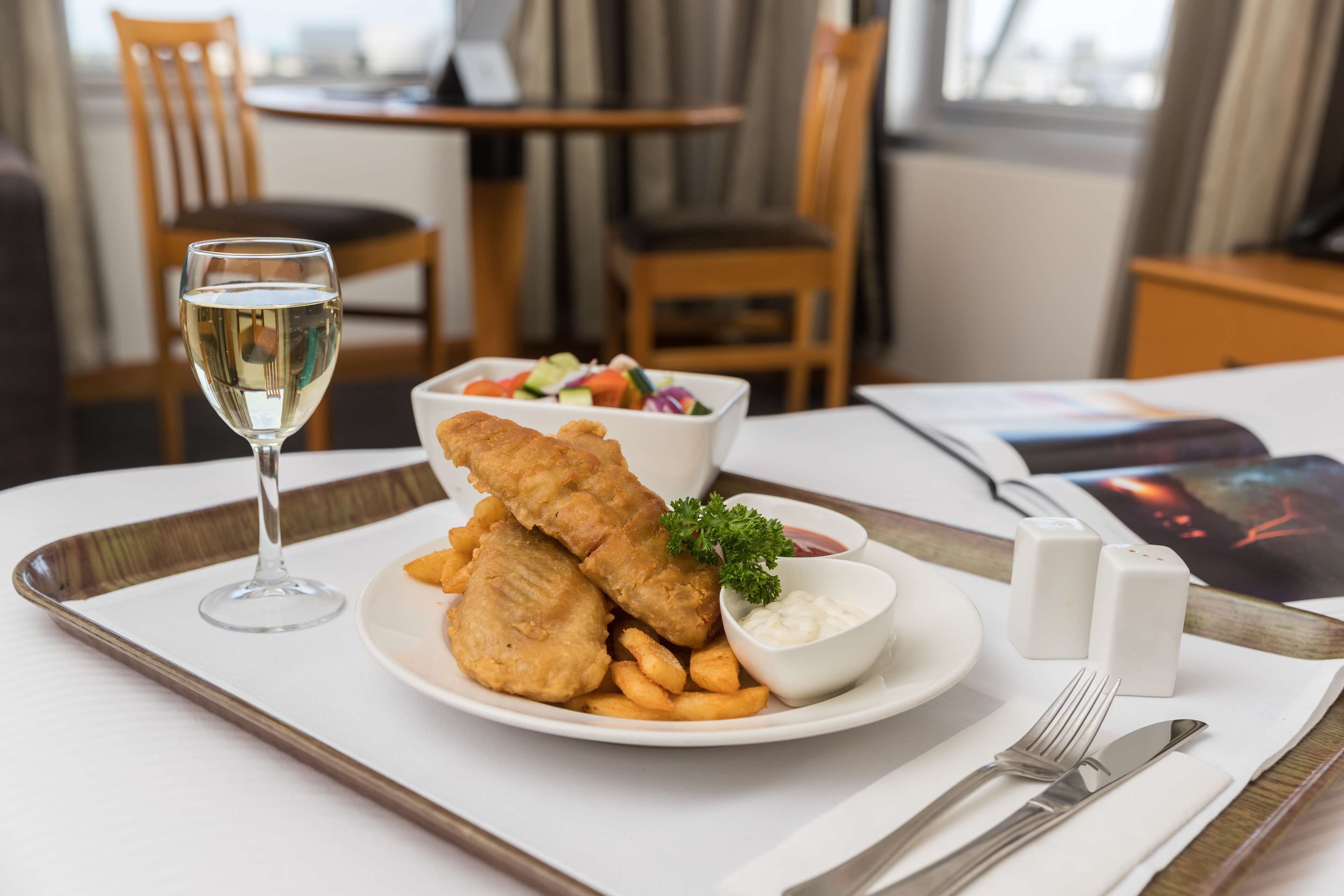 A dining table with a plate of fish and chips, a glass of white wine, and a small bowl of tartar sauce.