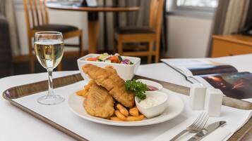 A dining table with a plate of fish and chips, a glass of white wine, and a small bowl of tartar sauce.