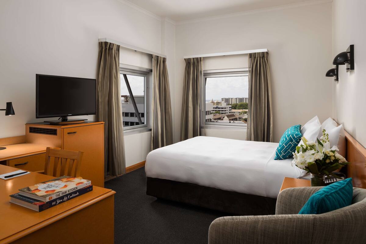 Habitación de hotel con cama, escritorio con televisor, una silla y vistas a edificios por las ventanas.
