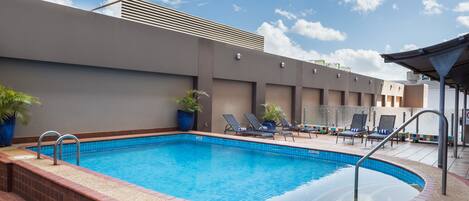 Una piscina con tumbonas y plantas en macetas, rodeada por un edificio moderno de techo plano y cielo despejado.