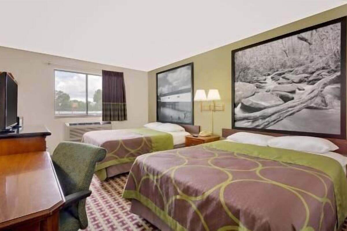 A hotel room with two beds, a desk, a chair, a window with curtains, and a large framed picture on the wall.