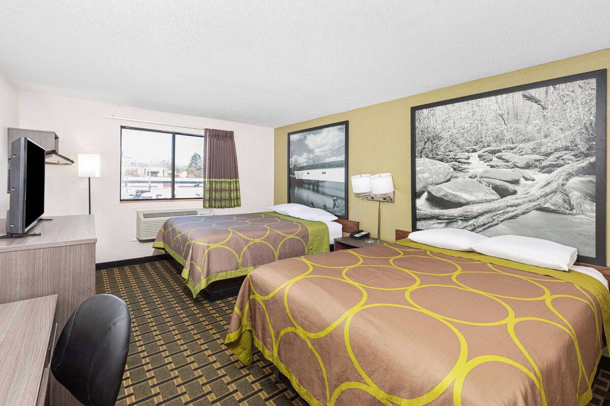 A hotel room with two beds, a desk, a TV, and a large window with curtains.