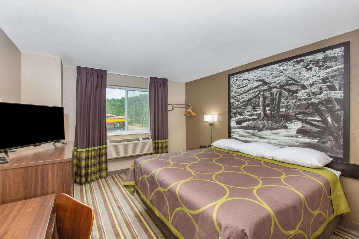 Habitación de hotel con cama, escritorio con televisor, ventana con cortinas, un cuadro en la pared y una lámpara.