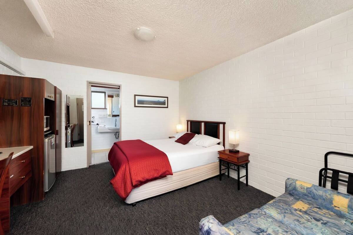 A hotel room with a bed, a desk, a chair, a small table with a lamp, and a picture on the wall.