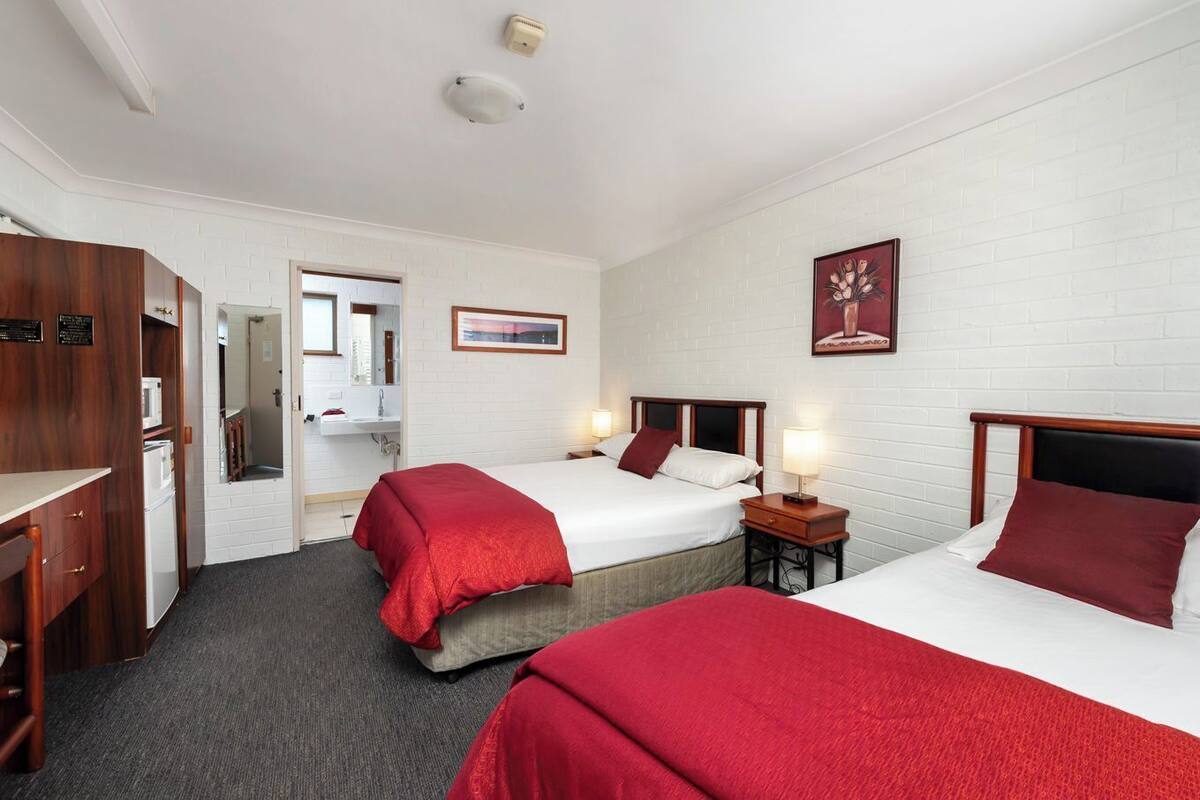 A hotel room with two beds, a desk, a wardrobe, a bathroom, and a white brick wall.