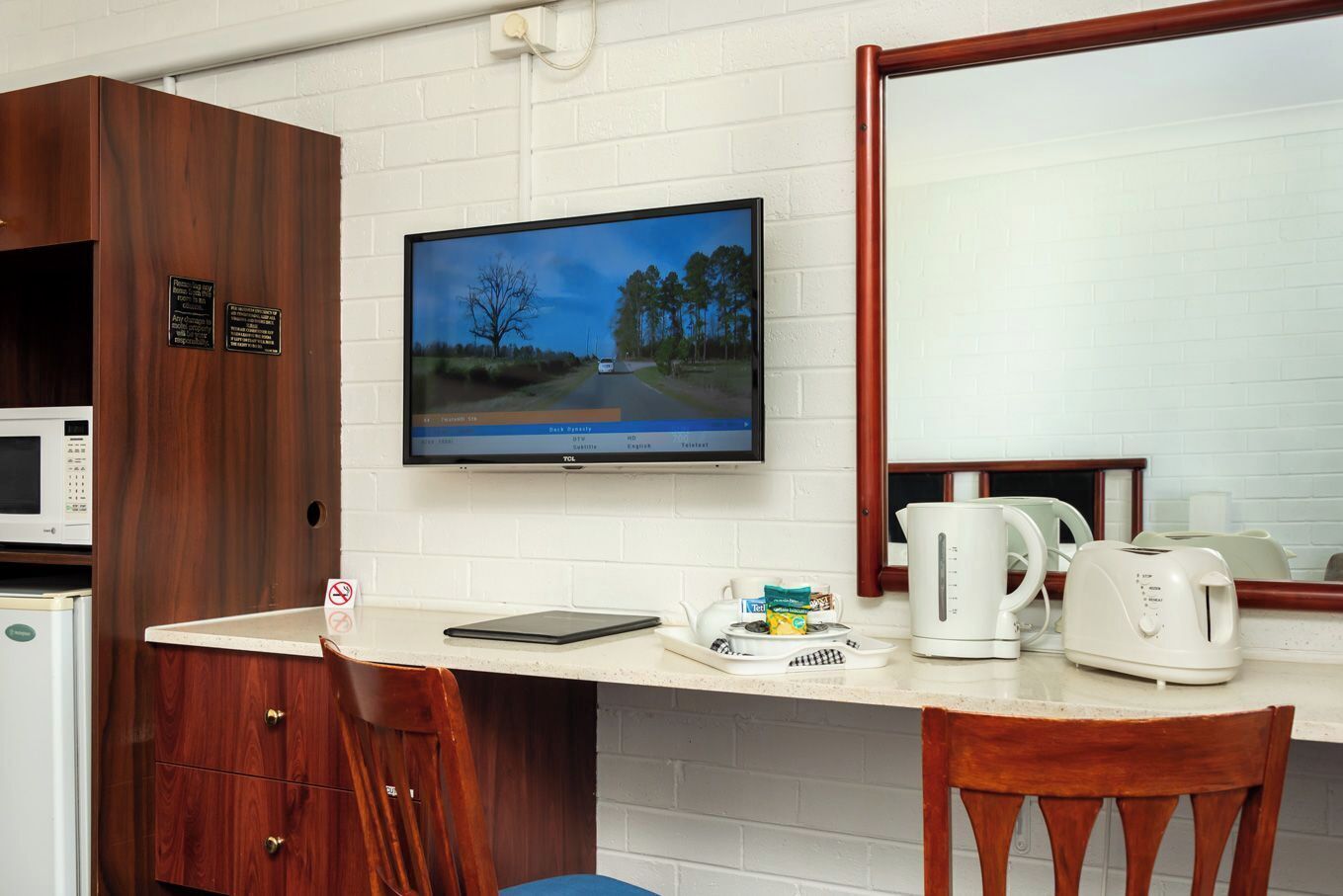 A kitchenette with a microwave, toaster, and kettle on the counter, a TV mounted on the wall, and a wooden cabinet with a sign.