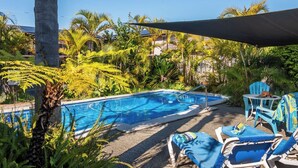 Piscina con sillas de descanso y sombrillas, rodeada de plantas tropicales y palmeras.