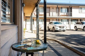 A glass table with a drink and a book, a balcony with a view of parked cars, and a building with multiple windows and balconies.
