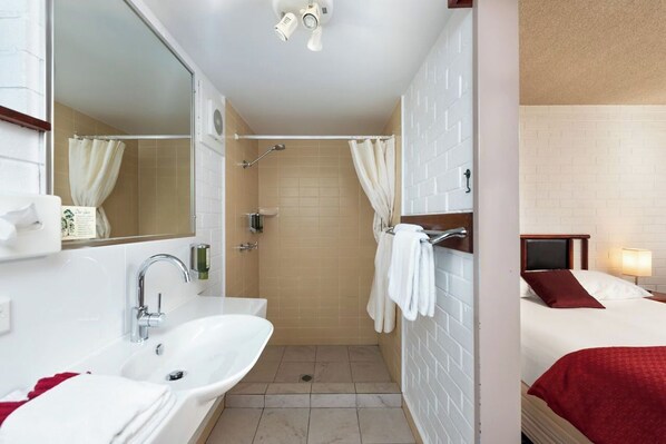A hotel bathroom with a sink, mirror, shower, and a bed with a red blanket.