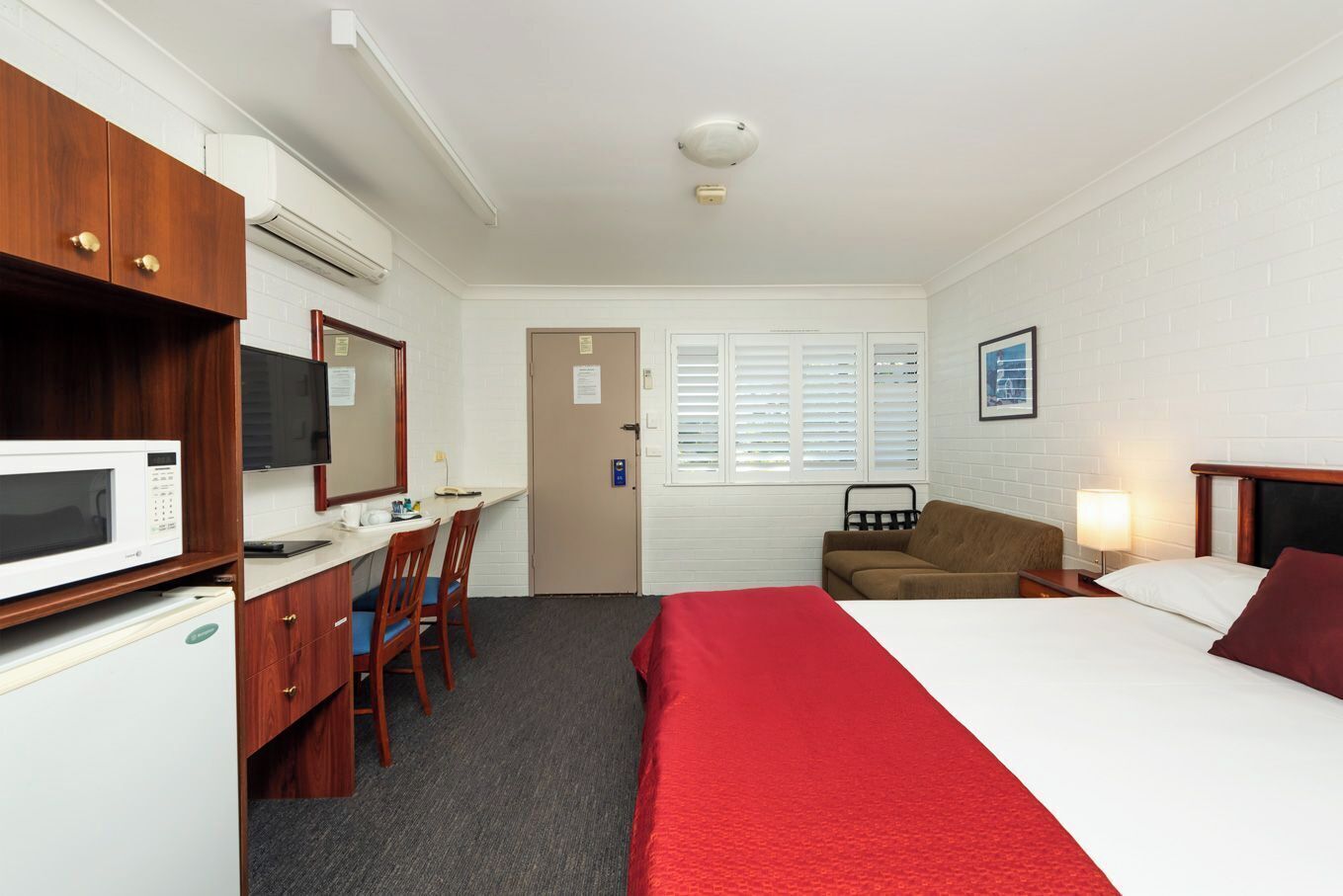 A hotel room with a bed, a desk with a microwave, a television, a sofa, and a window with blinds.