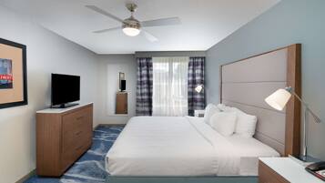 A hotel room with a bed, a TV, a desk, a lamp, a ceiling fan, and a window with curtains.
