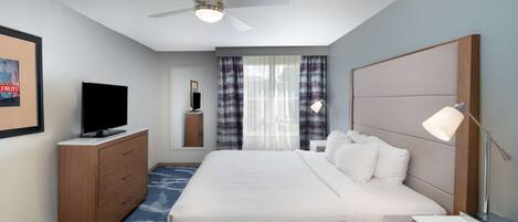 A hotel room with a bed, a TV, a desk, a lamp, a ceiling fan, and a window with curtains.