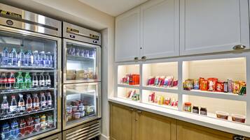 Mostrador refrigerado con diversas bebidas y una estantería con aperitivos y productos envasados.