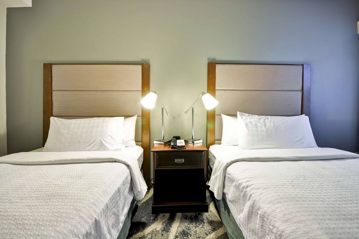 A hotel room with two beds, each with a wooden headboard, white bedding, and bedside lamps.