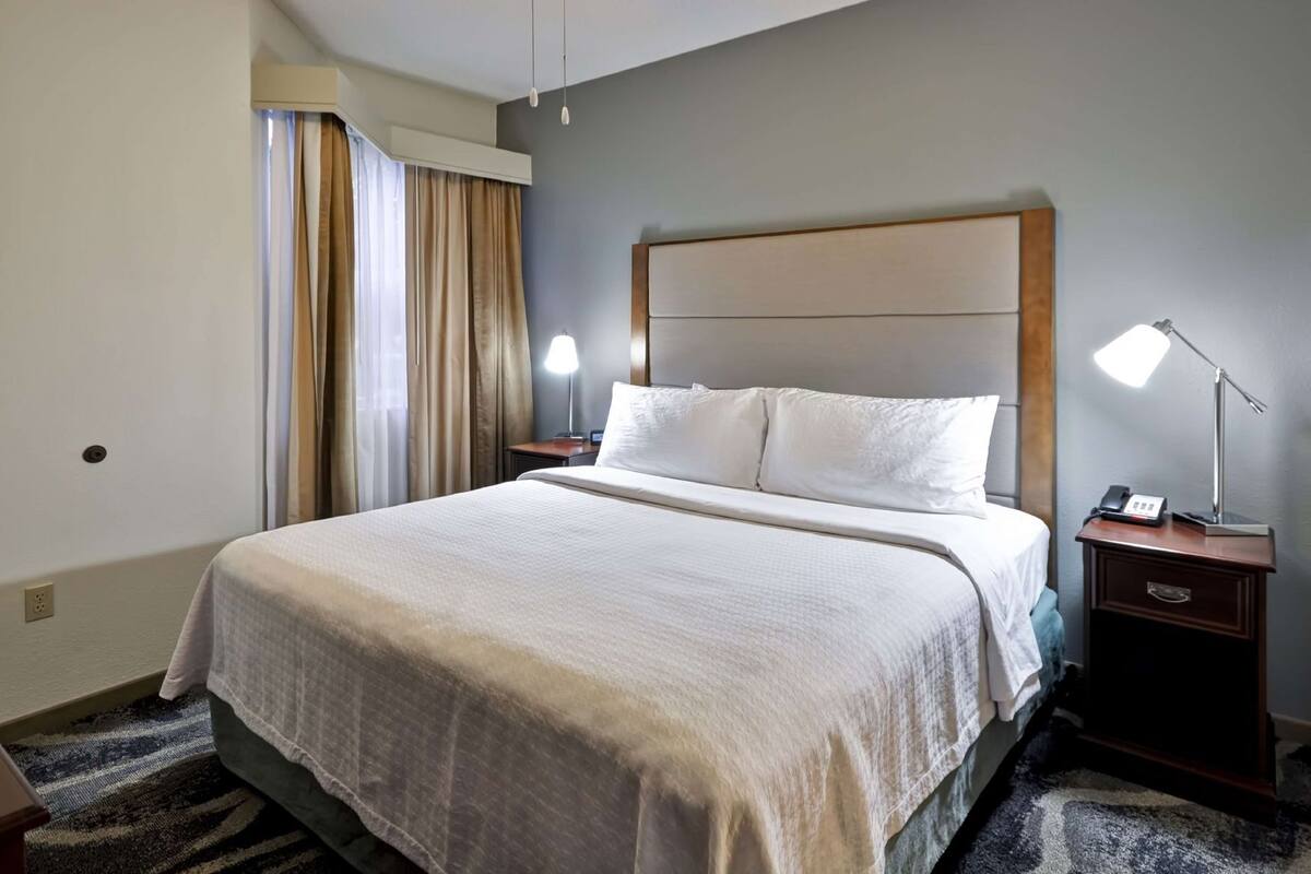 A hotel room with a large bed, two bedside lamps, a nightstand with a phone, and a window with curtains.