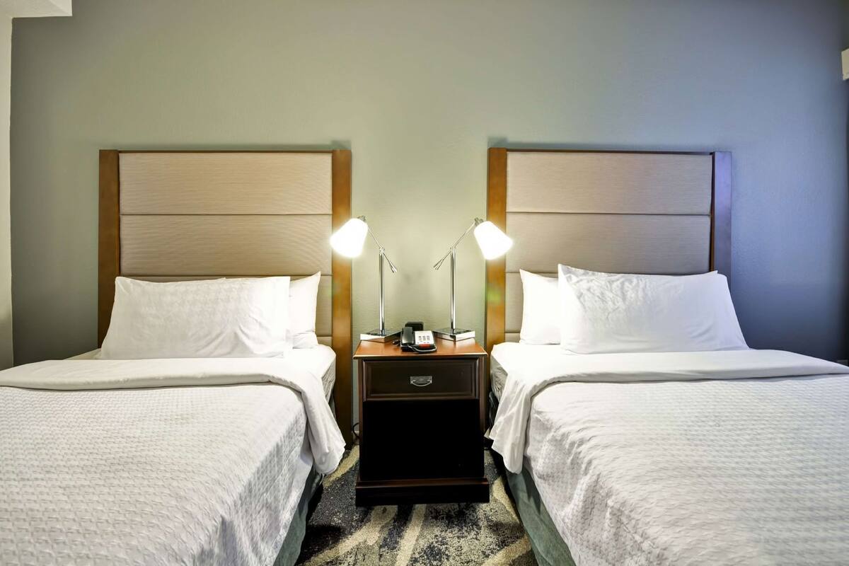 A hotel room with two beds, nightstands, lamps, and a small drawer.