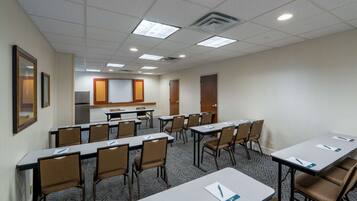 Sala de conferência com mesas e cadeiras, uma kitchenette ao fundo e uma porta à direita.