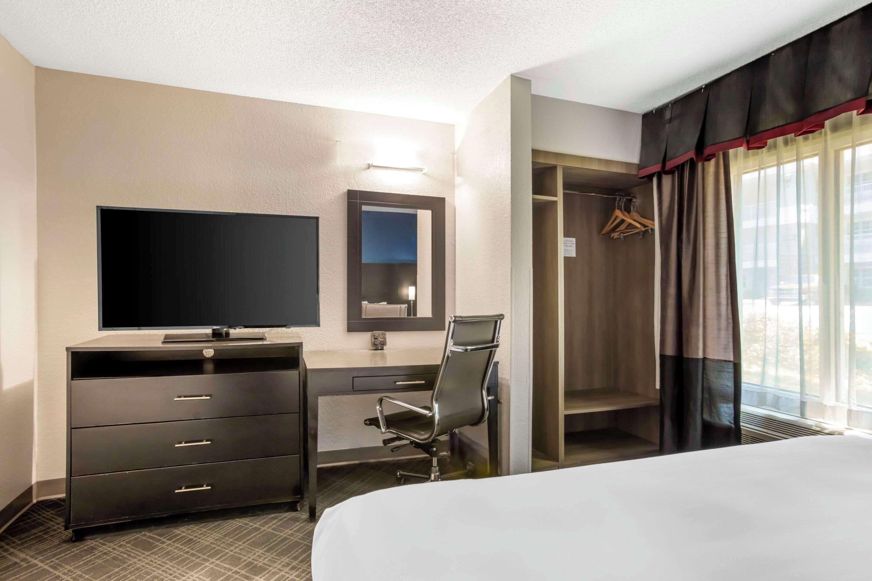 Une chambre d’hôtel comprenant un lit, un bureau avec une chaise, une télévision, un miroir, une armoire et une fenêtre avec des rideaux.