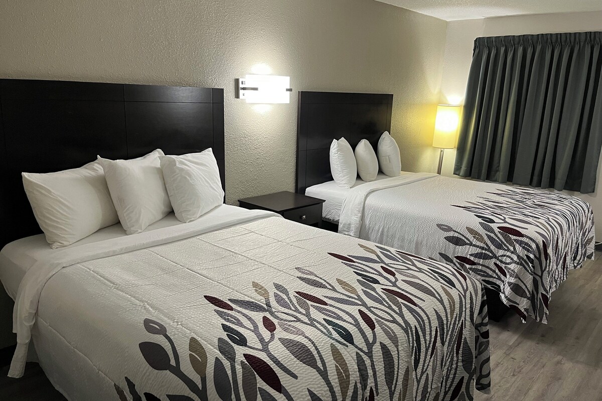 A hotel room with two beds, each with white bedding and a leaf-patterned duvet cover. There is a nightstand with a lamp between the beds, and a window with curtains.