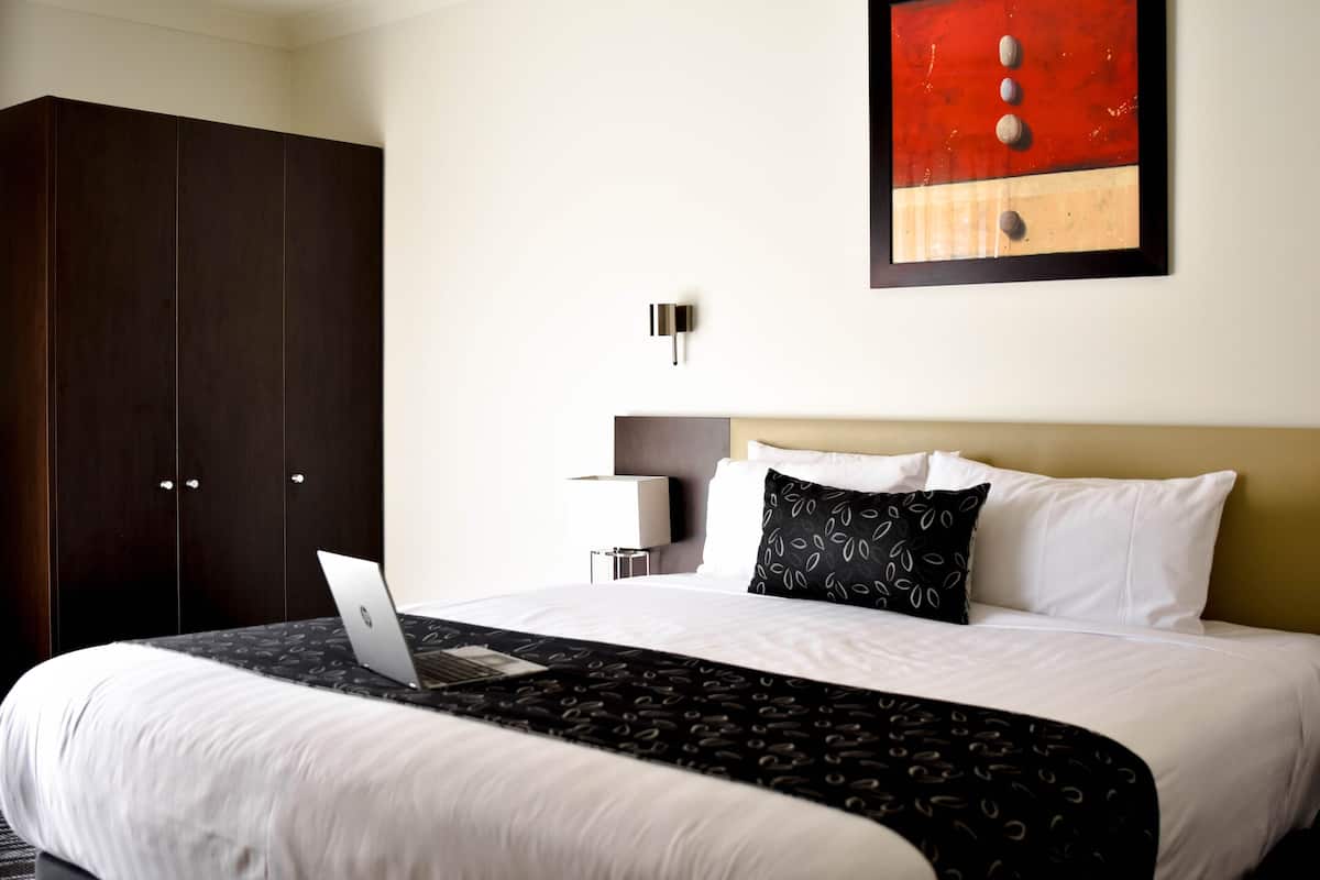 A hotel room with a bed, a laptop on the bed, a wardrobe, a wall-mounted light, and a painting.