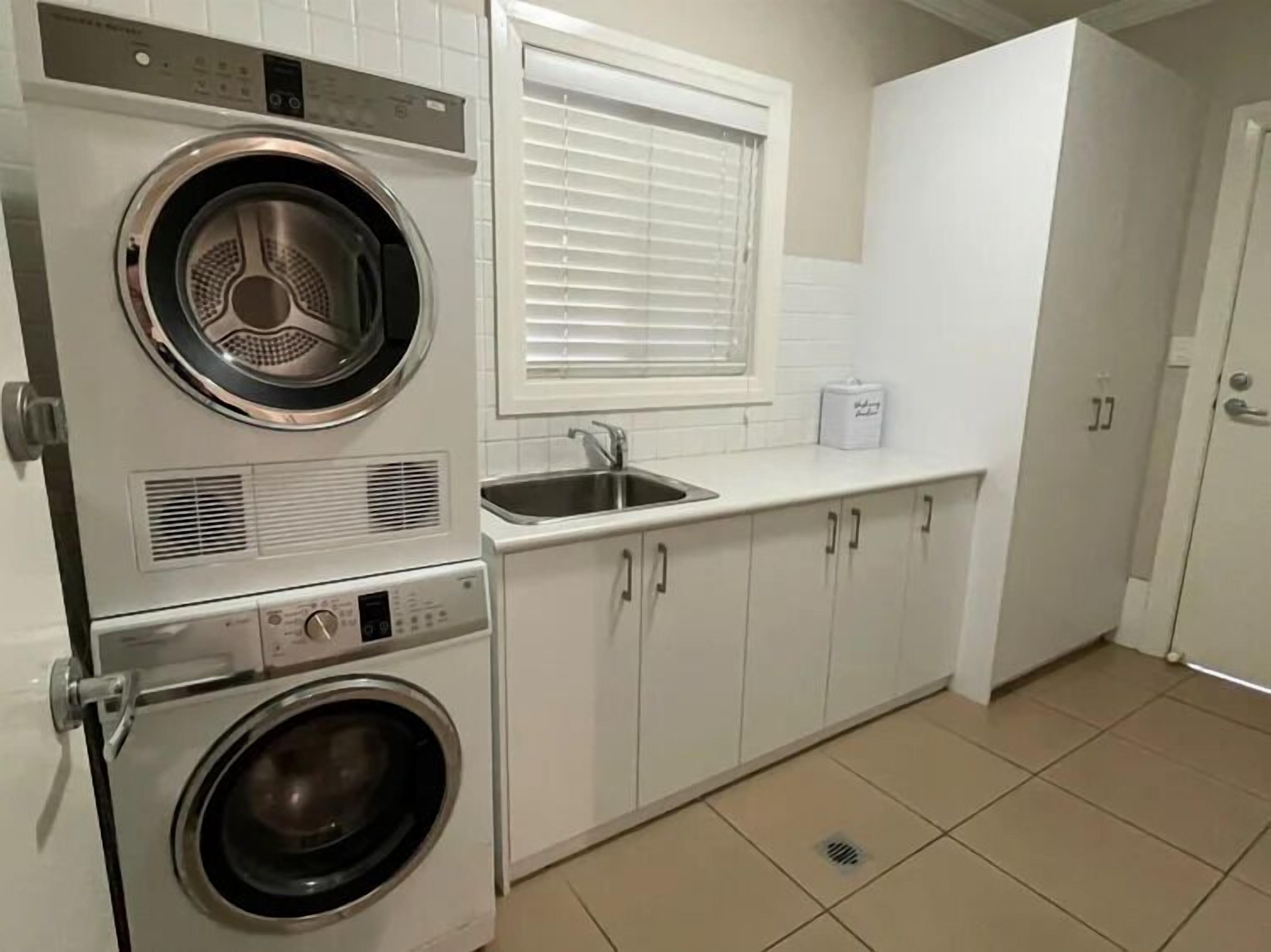 A laundry room with stacked washers and dryers, a sink, and a window with blinds.