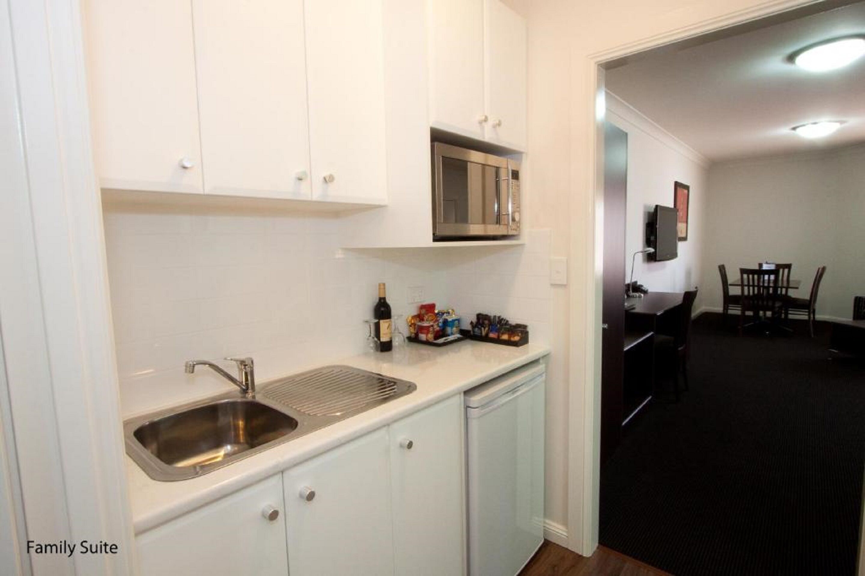 Una cocina con armarios blancos, un lavamanos de acero inoxidable, un microondas y una botella de vino. Se observa un comedor con una mesa y sillas al fondo.