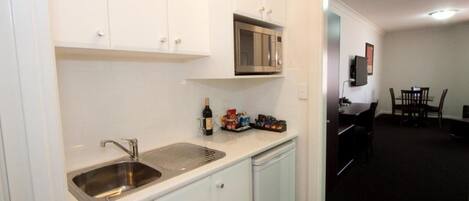 Una cocina con armarios blancos, un lavamanos de acero inoxidable, un microondas y una botella de vino. Se observa un comedor con una mesa y sillas al fondo.