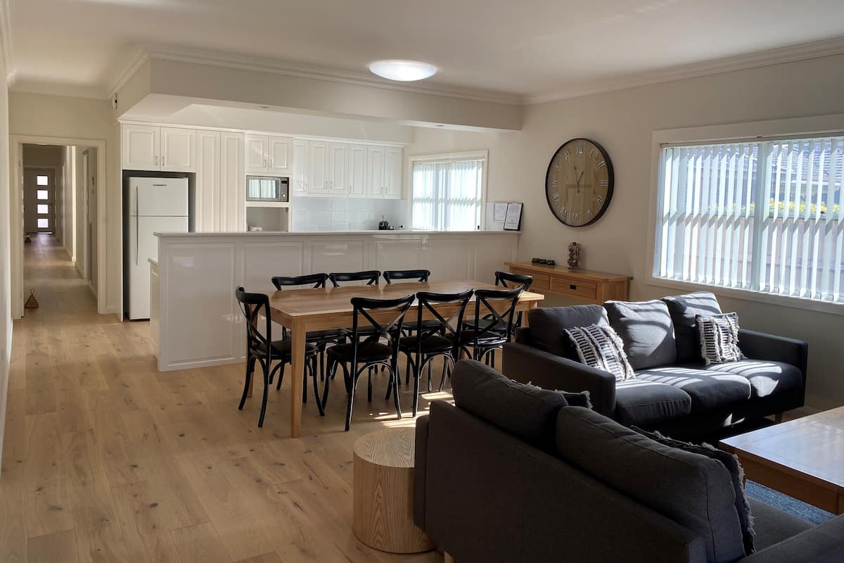 A modern kitchen with white cabinets, a wooden dining table, and black chairs. There is a large clock on the wall and a window with blinds.