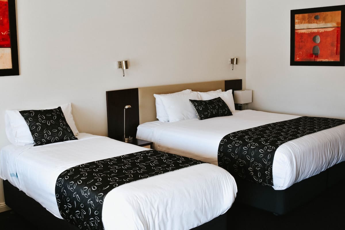 A hotel room with two beds, each with white linens and black patterned bedspreads. There are bedside tables with lamps and a wall-mounted light fixture.
