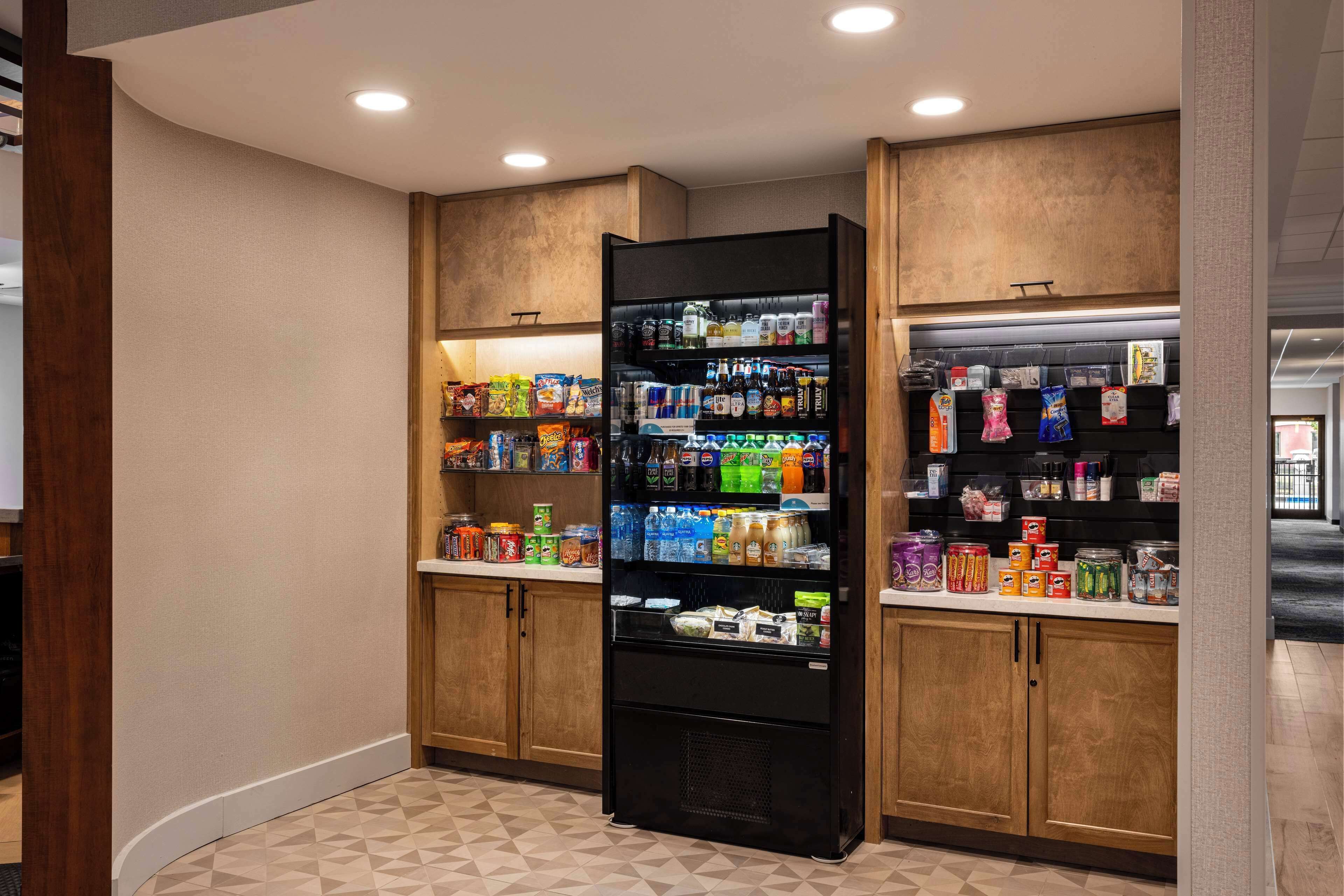 A well-lit hallway with a vending machine stocked with snacks and beverages, wooden cabinets, and a patterned floor.
