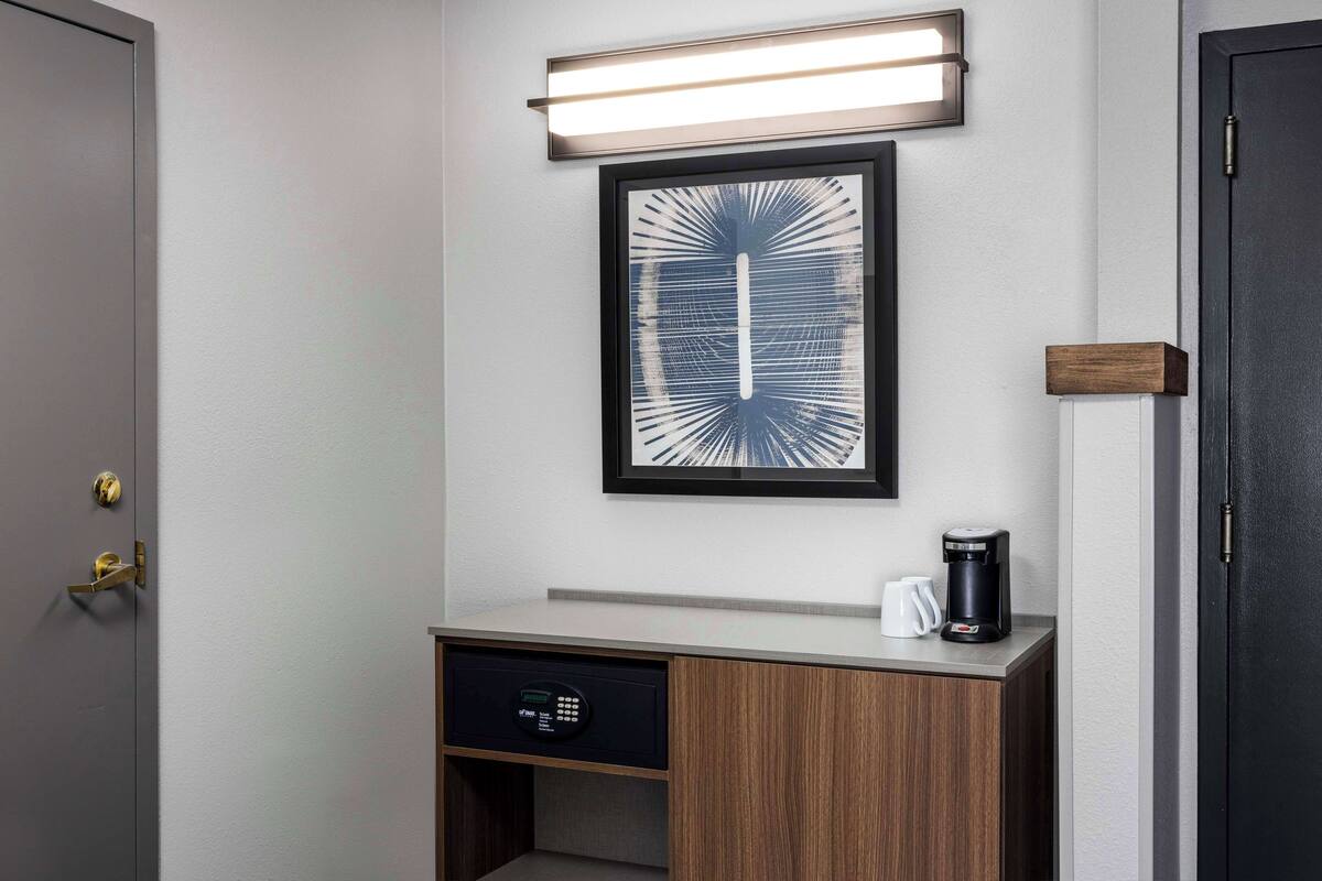 Habitación de hotel con una obra de arte enmarcada, una cafetera y un panel de control en un mueble de madera.