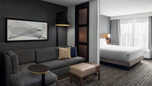 A hotel room with a grey sofa, a round table, a bed with white linens, a large abstract painting, a floor lamp, and a window with curtains.
