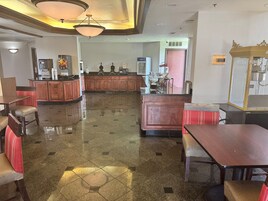Un vestíbulo de hotel con una recepción central, un comedor con mesas y sillas, y una cocina al fondo.