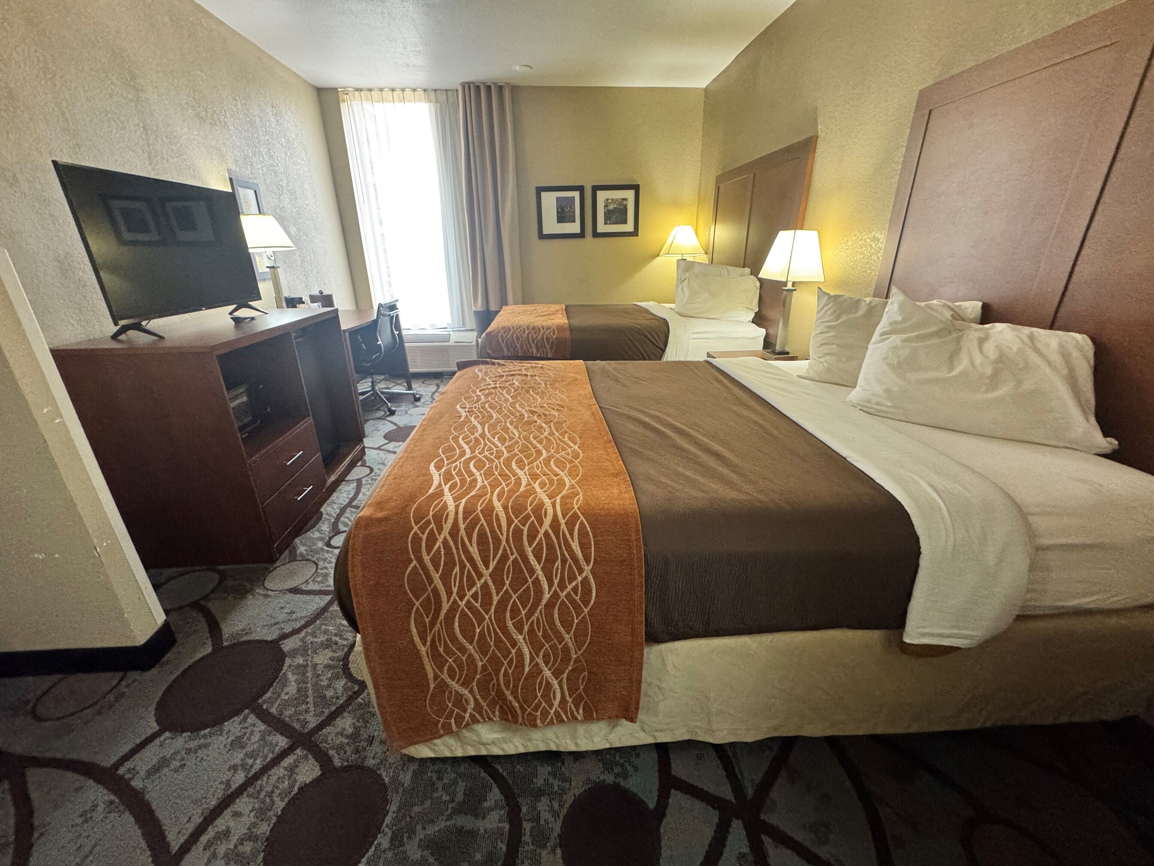 A hotel room with a bed, a desk with a computer, a TV, two bedside lamps, and a window with curtains.