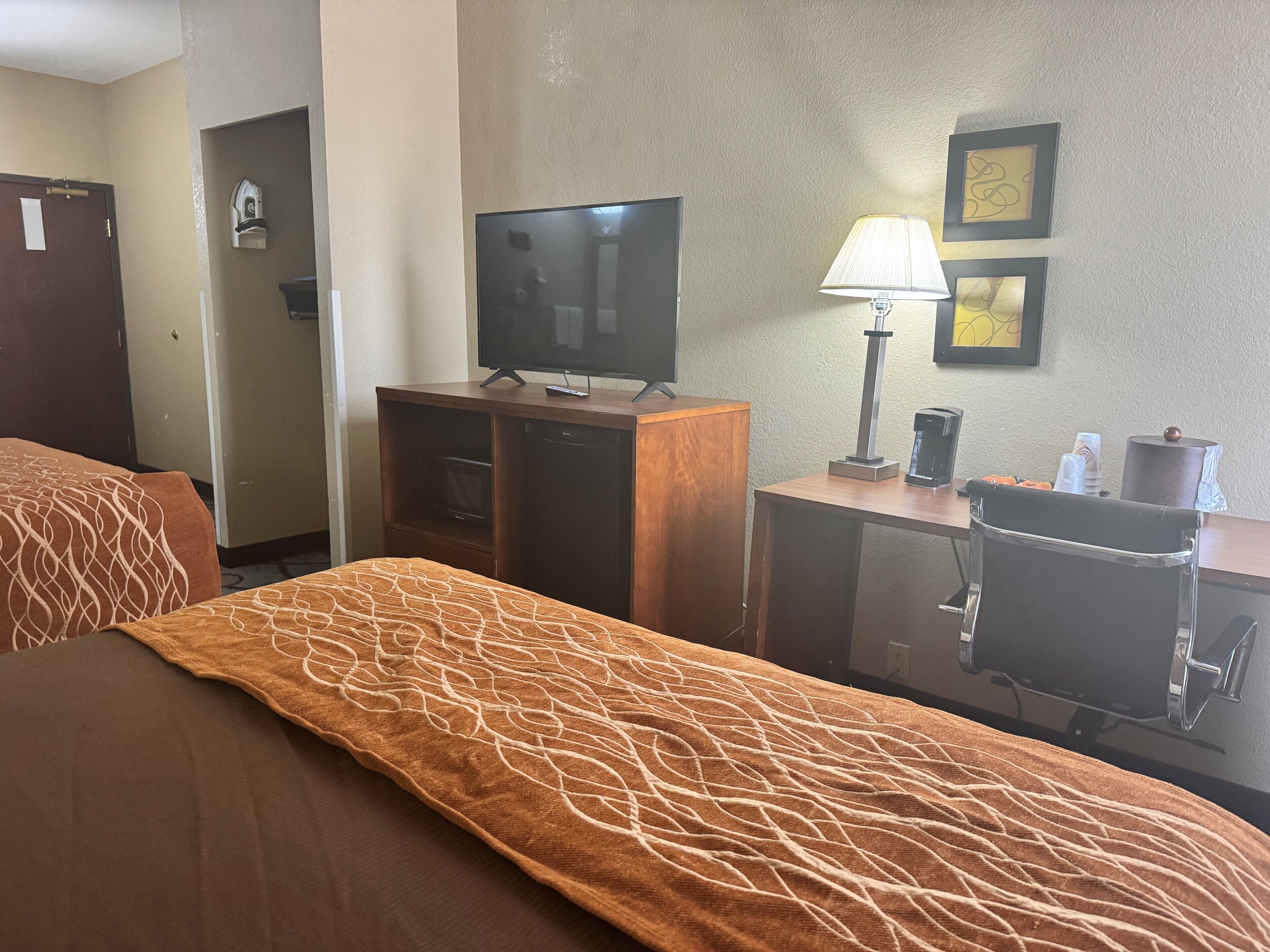 A hotel room with a bed, a TV on a wooden stand, a desk with a lamp, and a coffee machine.