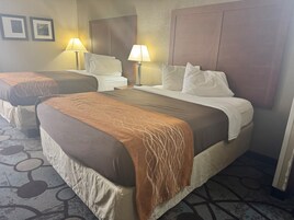 A hotel room with two beds, each with a brown headboard, white linens, and a patterned throw. There are two bedside lamps and a large mirror above the beds.