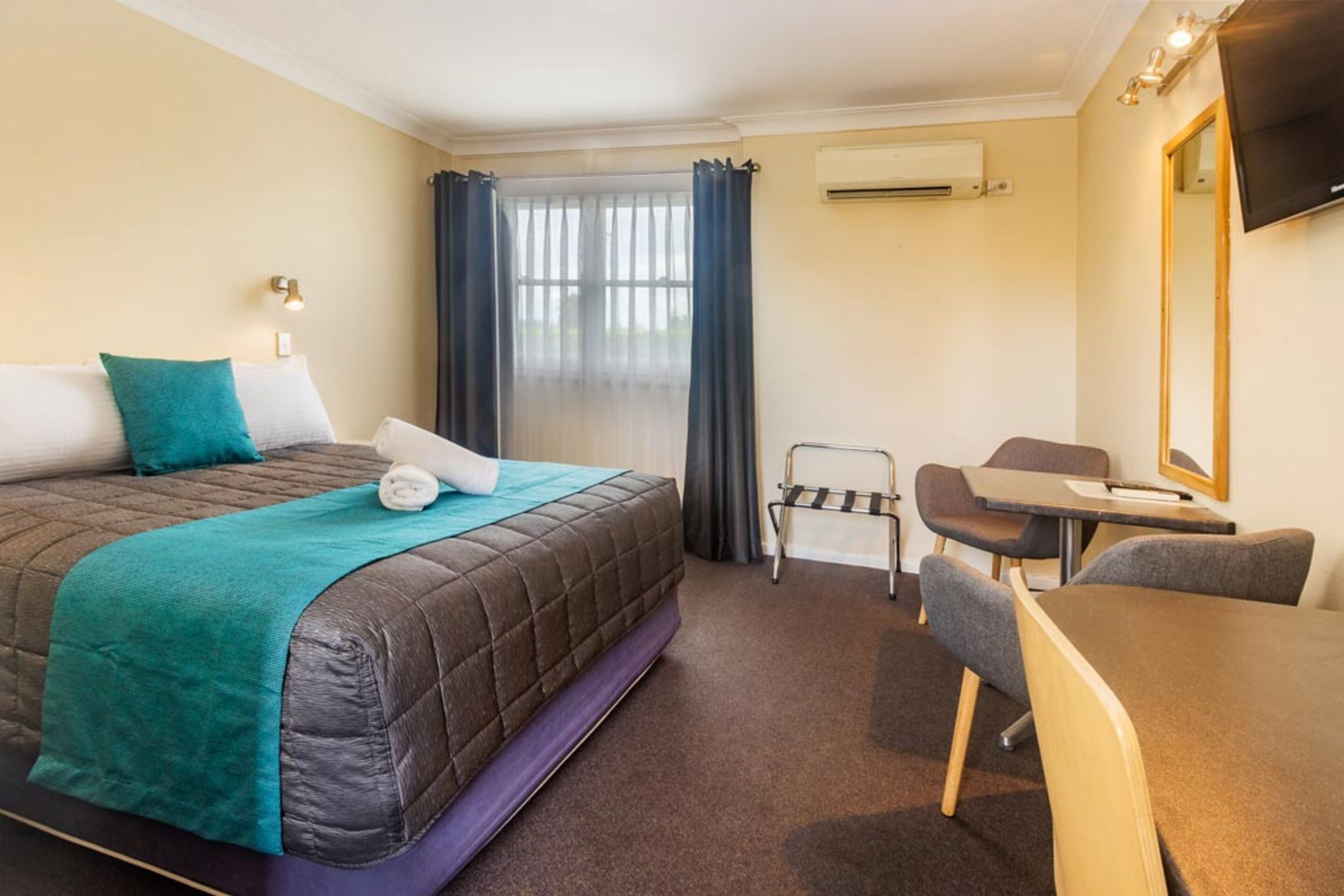 Quarto de hotel com cama, escrivaninha, cadeira, espelho e janela com cortinas.