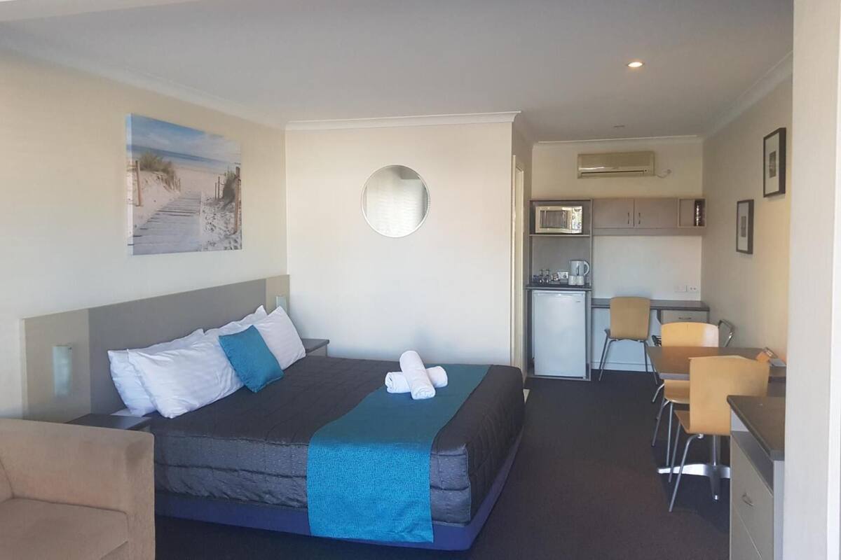 A hotel room with a bed, a desk, a chair, a small kitchenette, and a wall-mounted mirror.