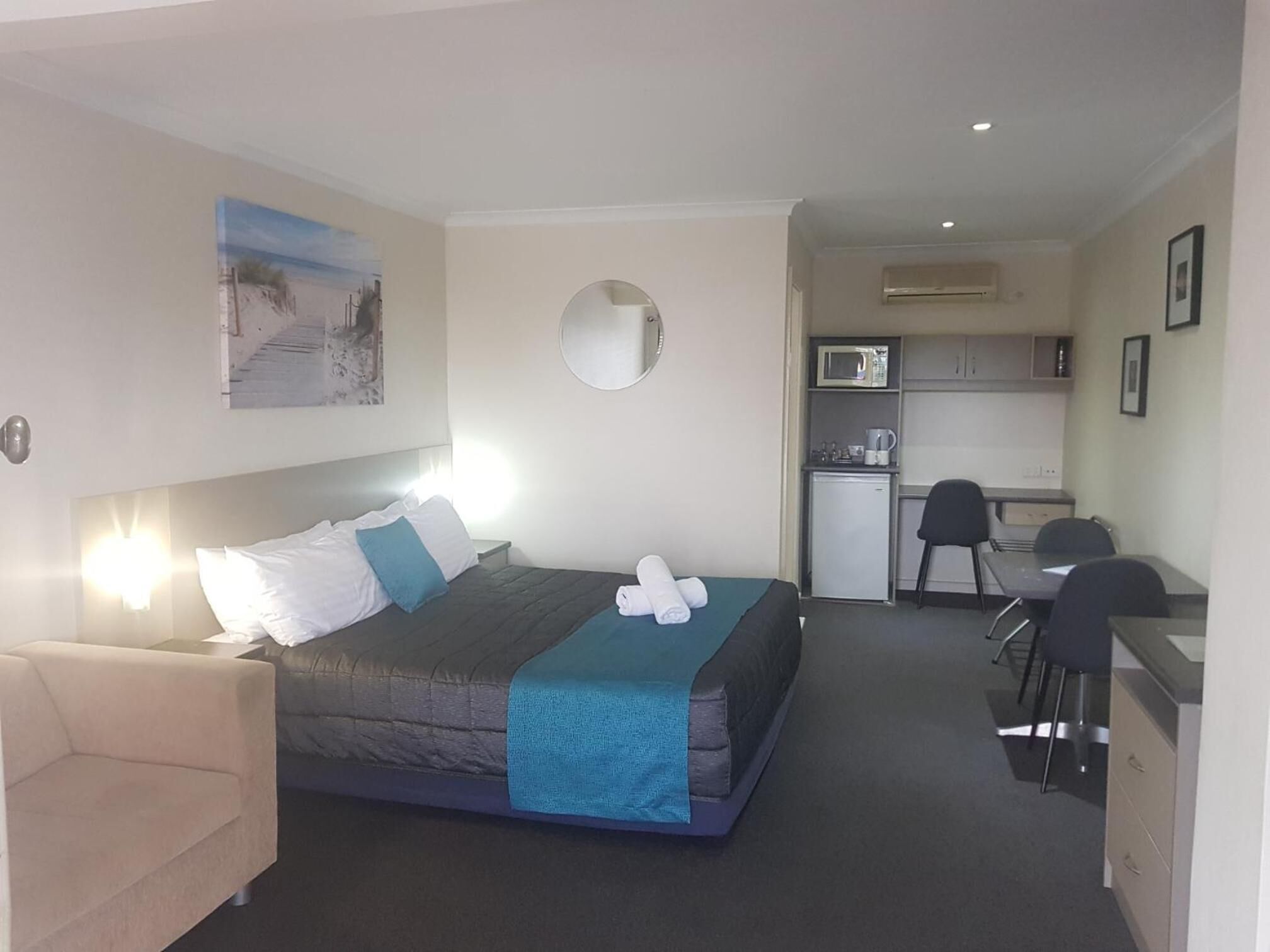 A hotel room with a bed, a desk, a chair, a small kitchenette, and a mirror.