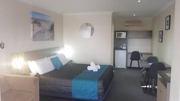 A hotel room with a bed, a desk, a chair, a small kitchenette, and a mirror.